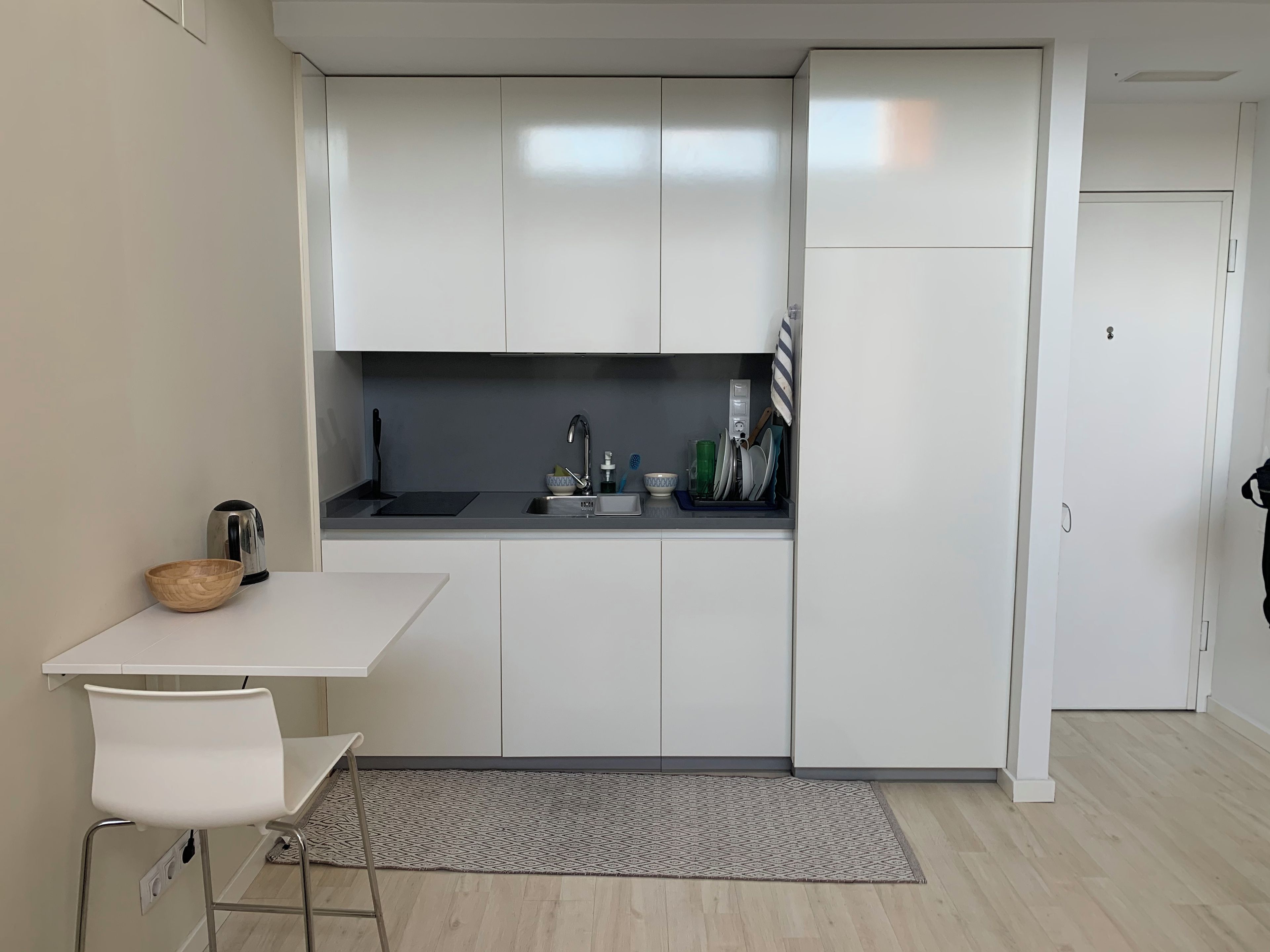 Une cuisine compacte avec des meubles blancs, un évier, une bouilloire et une petite table à manger avec une chaise.
