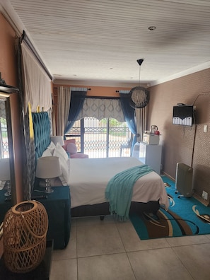 A hotel room with a bed, a desk, a chair, a lamp, a hanging light fixture, and a view of the outdoors. - Room-i am a Dedicate, Reliable and Hardw (Matjhabeng)