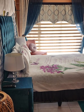 A bedroom with a bed, a nightstand with a lamp, a blue upholstered headboard, and a window with curtains. - Room-i am a Dedicate, Reliable and Hardw (Matjhabeng)