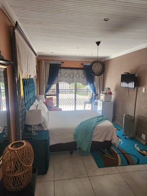 A hotel room with a bed, a desk, a chair, a lamp, a hanging light fixture, and a view of the outdoors. - Room-i am a Dedicate, Reliable and Hardw (Matjhabeng)