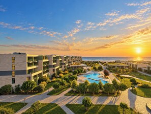 Un hotel moderno con piscina, senderos en un jardín y vista al océano al atardecer.