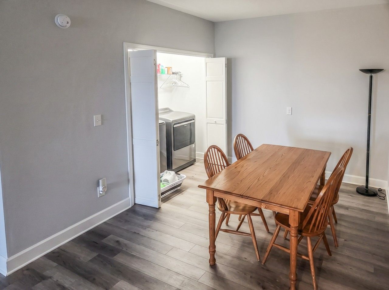 Un comedor con una mesa y sillas de madera, una cocina al fondo y una lámpara de pie.