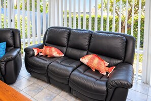 Un sofá de cuero negro con dos almohadas con forma de pez, una almohada azul y una mesa de madera en una habitación soleada con vistas a una cerca blanca y follaje.