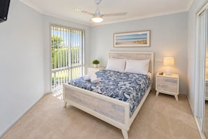 Quarto com cama, ventoinha no teto, mesa de cabeceira, abajur e janela com persianas.