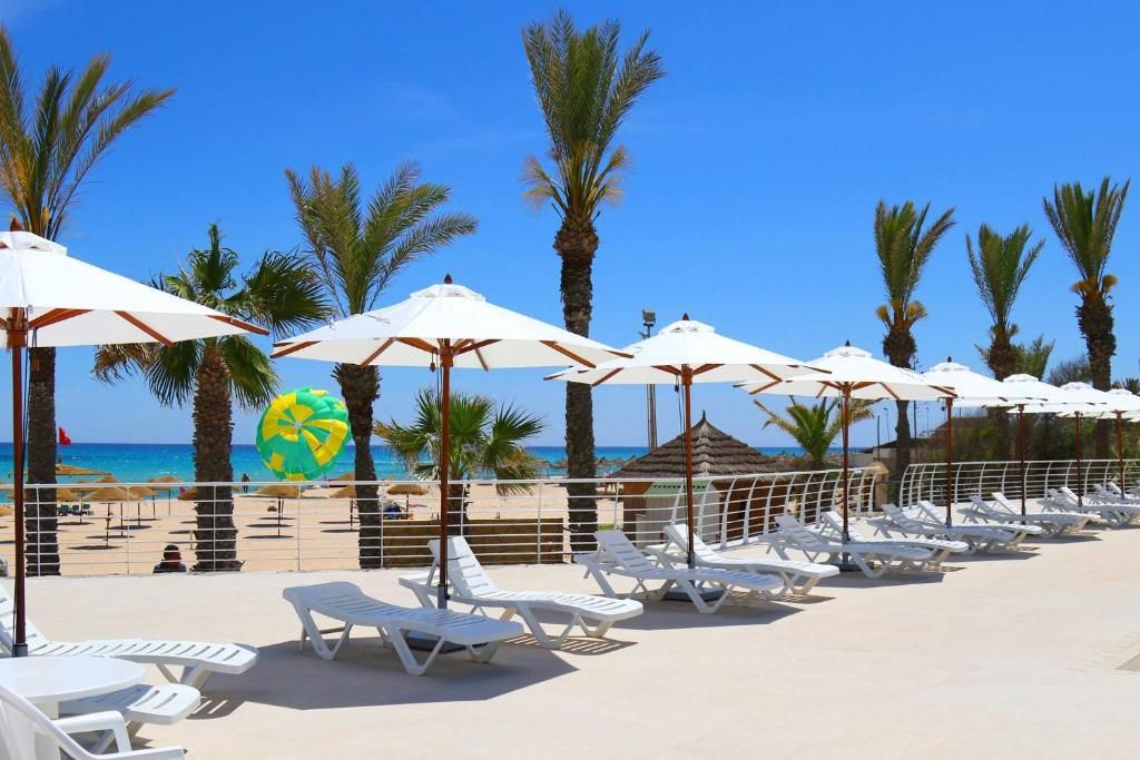 A beachside lounge area with sun loungers and umbrellas, palm trees, and a clear view of the sea.