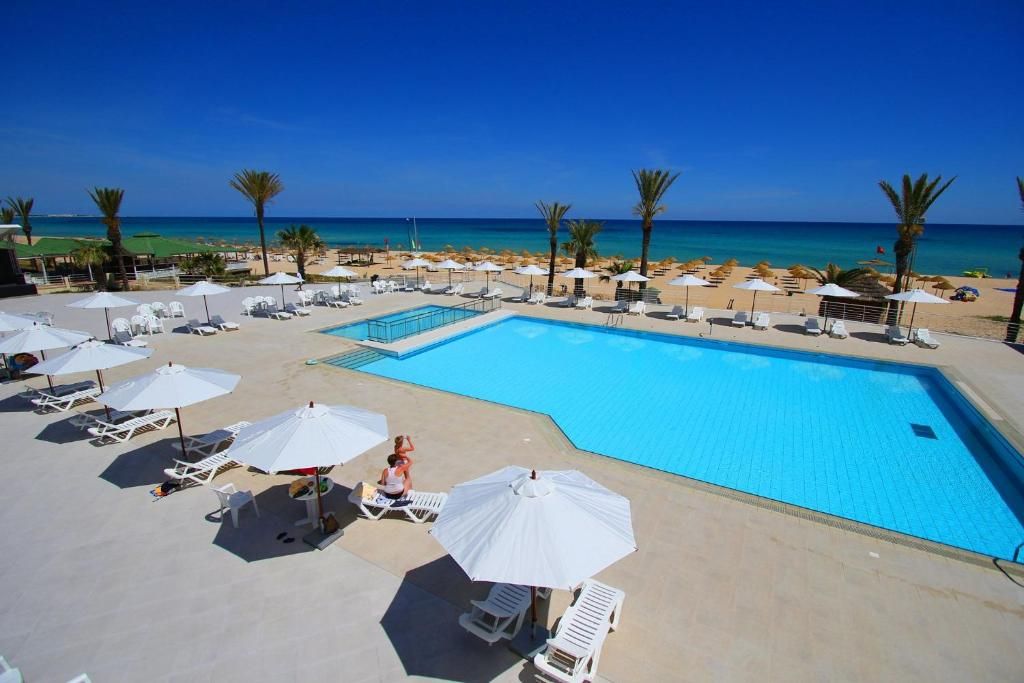 Un espace piscine avec des chaises longues et des parasols, un ciel d’un bleu éclatant et une plage en arrière-plan.