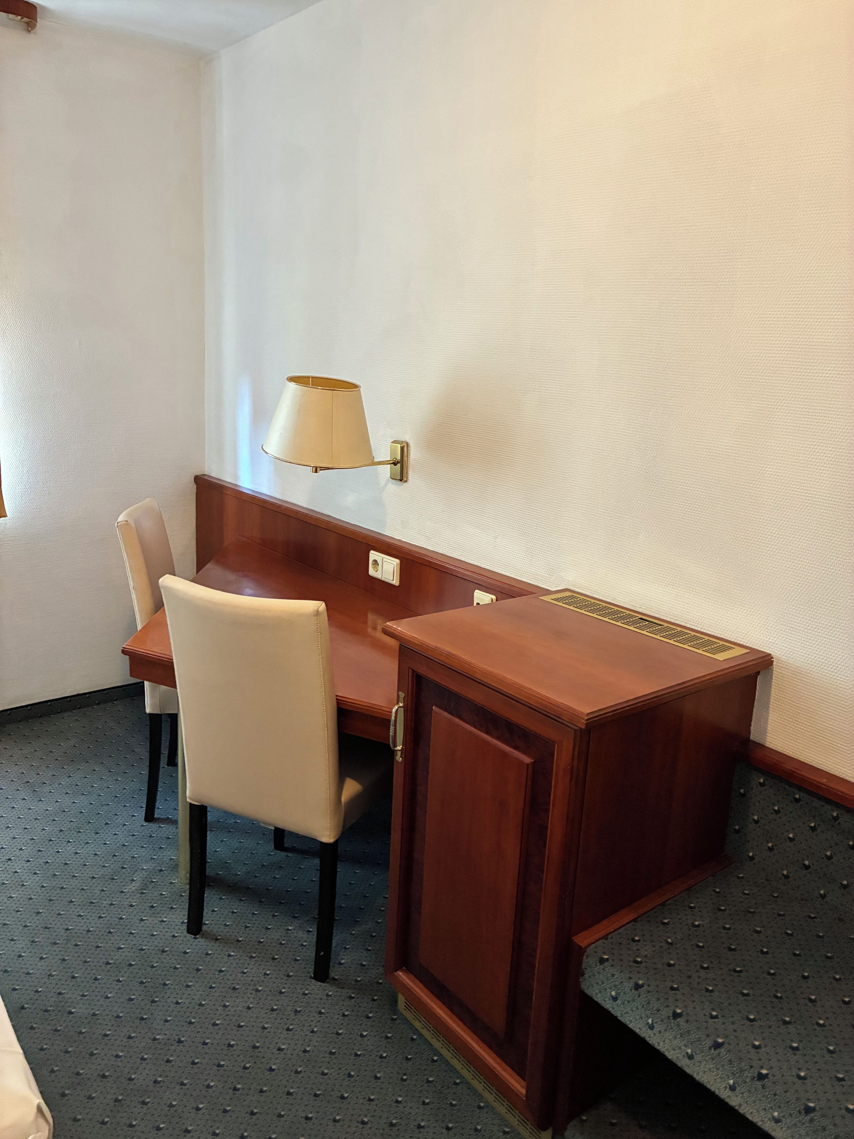 A hotel room with a wooden desk, a chair, a lamp, and a bed.