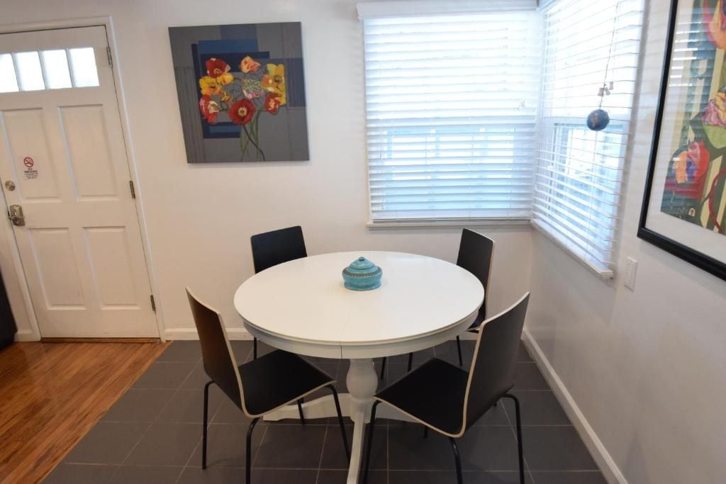 A dining area with a round table and four chairs, a painting on the wall, and a window with blinds.