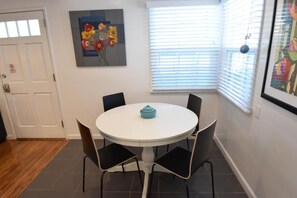 Uma sala de jantar com uma mesa redonda e quatro cadeiras, um quadro na parede e uma janela com persianas.