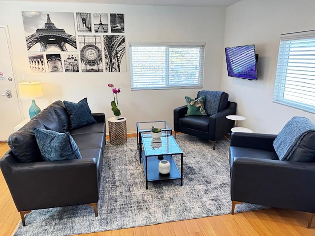 A living room with two black sofas, a glass coffee table, and a rug. There are wall art prints, a TV, and a window with blinds.
