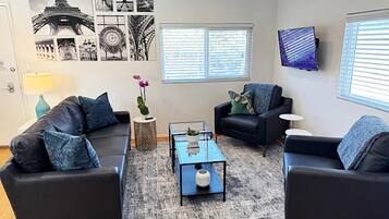 A living room with two black sofas, a glass coffee table, and a rug. There are wall art prints, a TV, and a window with blinds.