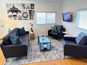 Uma sala de estar com dois sofás pretos, uma mesa de centro de vidro e um tapete. Há quadros nas paredes, uma televisão e uma janela com persianas.