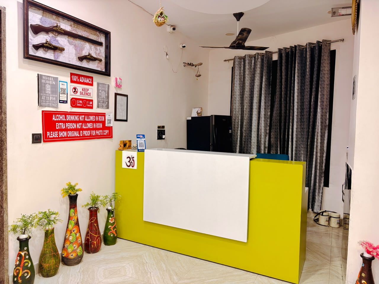 A hotel reception desk with a yellow counter, a white towel, and several vases with plants. There are signs on the wall, including one about alcohol not being allowed in the room.