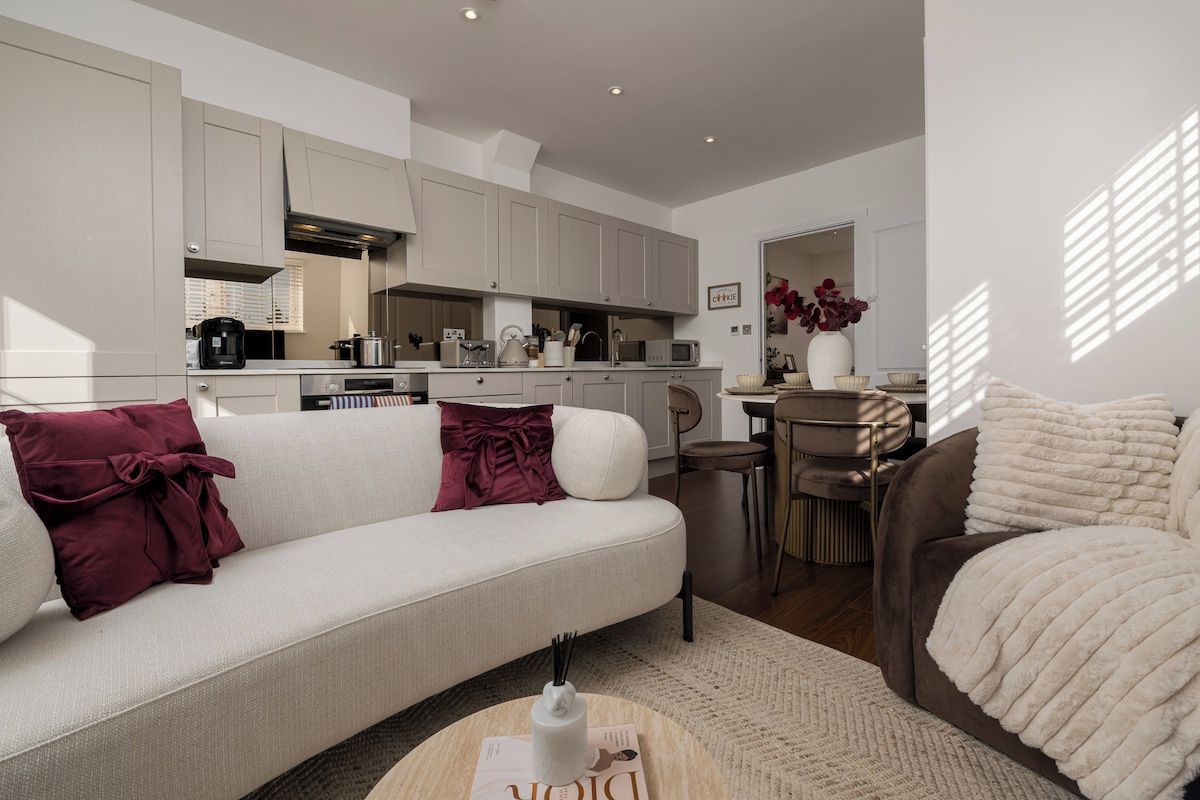 A modern living room with a sofa, a coffee table, and a brown armchair. There is a kitchen in the background with cabinets and appliances.