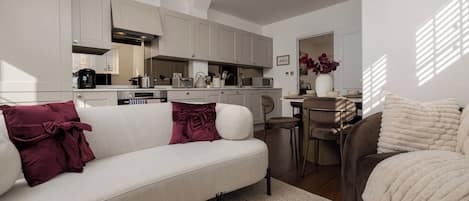 A modern living room with a sofa, a coffee table, and a brown armchair. There is a kitchen in the background with cabinets and appliances.