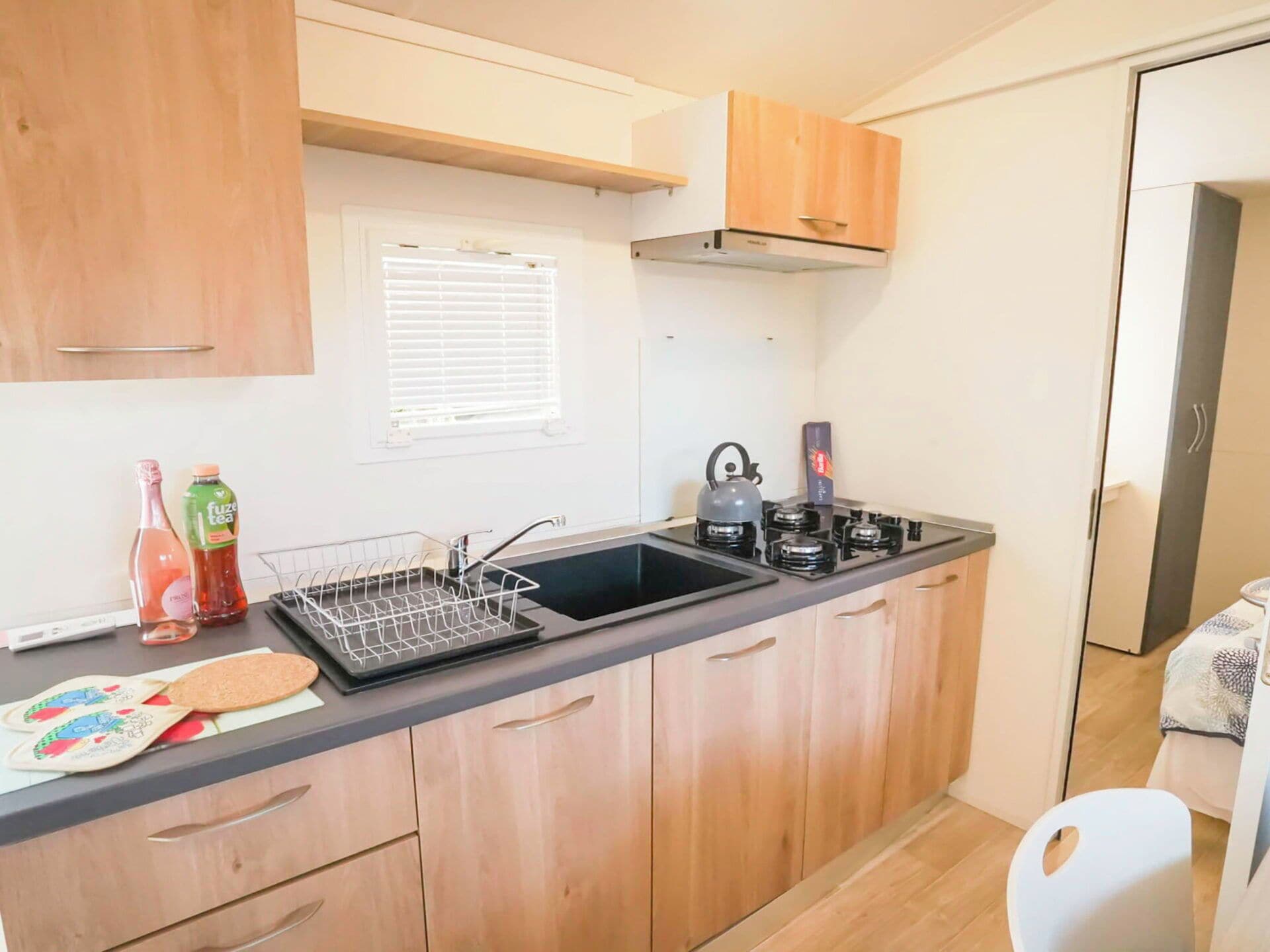 Una kitchenette compacta con un lavabo negro, una tetera y una cafetera. Hay armarios de madera y una ventana con persianas.