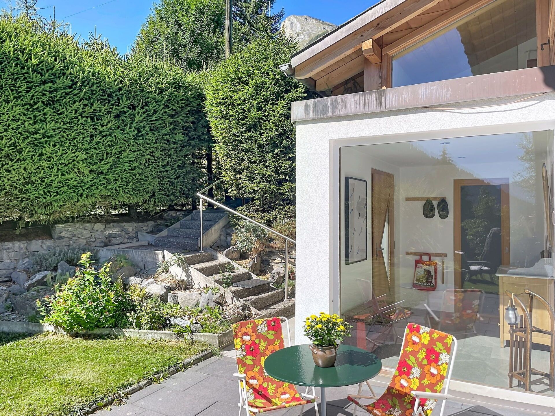 A modern outdoor seating area with a glass door leading to a room, surrounded by a stone wall and a well-maintained garden.