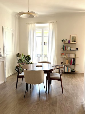 Ein Essbereich mit einem runden Tisch, vier Stühlen, einer Hängelampe, einem Bücherregal und einem Fenster mit Vorhängen.