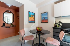 Een moderne keuken met een zwart aanrecht, een spoelbak en een koffieapparaat. Er staan twee roze stoelen bij een ronde tafel met een schaal fruit.
