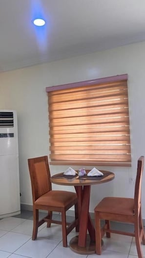 A dining area with a round table, two chairs, and a window with blinds. - Nbte hotels (Kaduna/kaduna state)