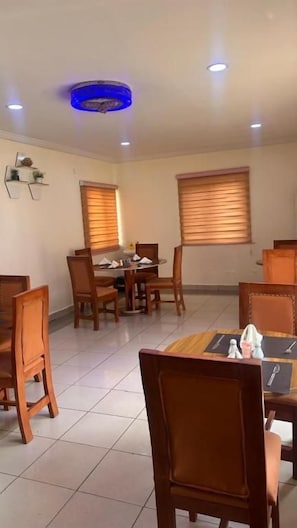 A dining area with a round table and chairs, a ceiling light fixture, and windows with blinds. - Nbte hotels (Kaduna/kaduna state)