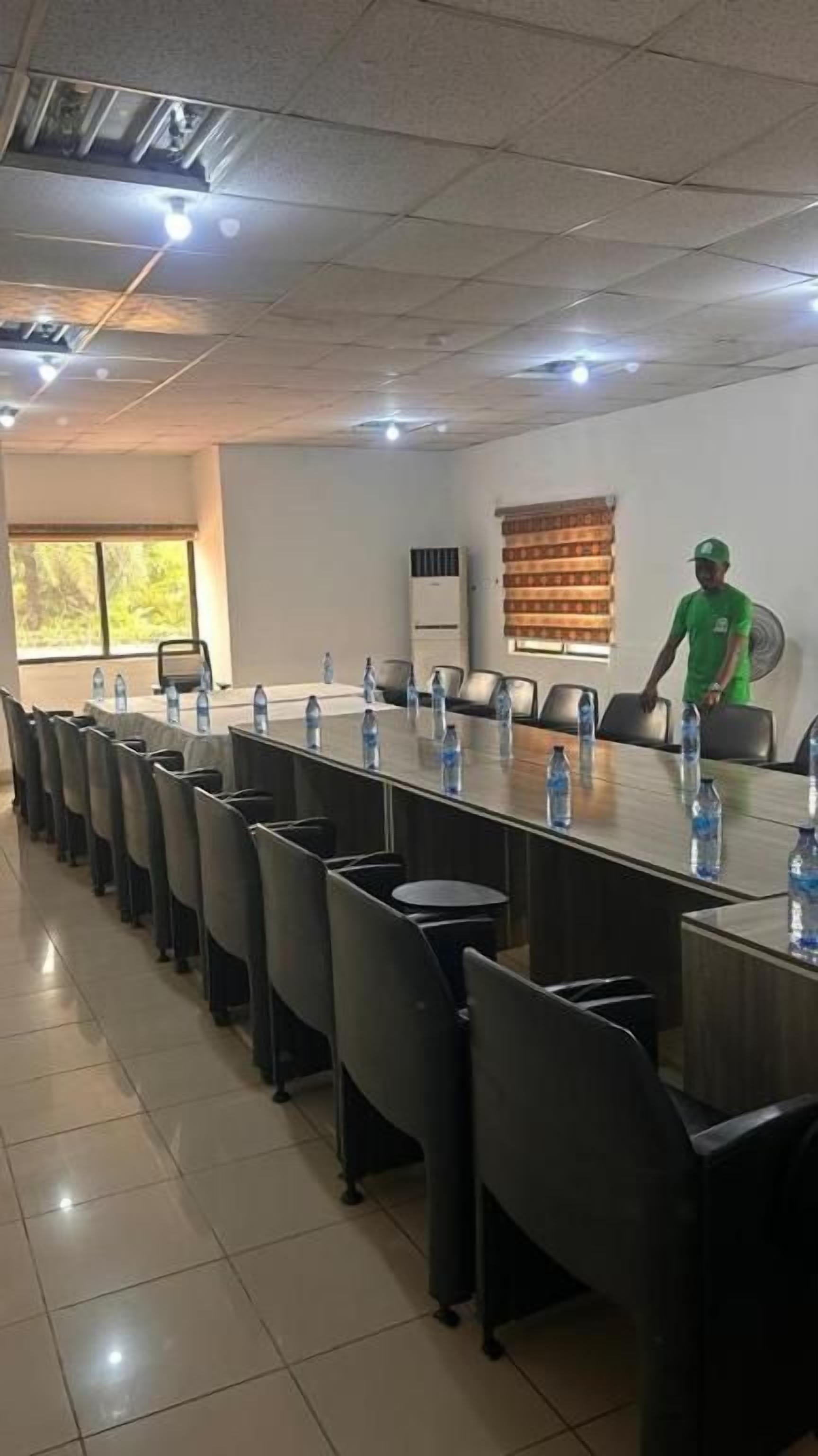 A conference room with a long table, chairs, water bottles, and a person in a green shirt standing by the window.