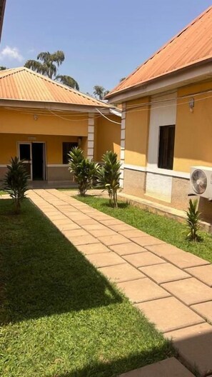 Une cour avec un chemin pavé, de petits palmiers et un bâtiment doté d’un toit en tuiles.