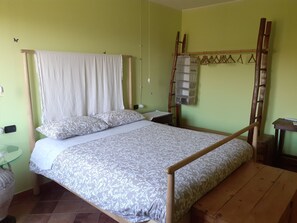 Un lit simple avec un cadre en bois et un baldaquin, une table de chevet, une étagère fixée au mur et une petite table avec une lampe.