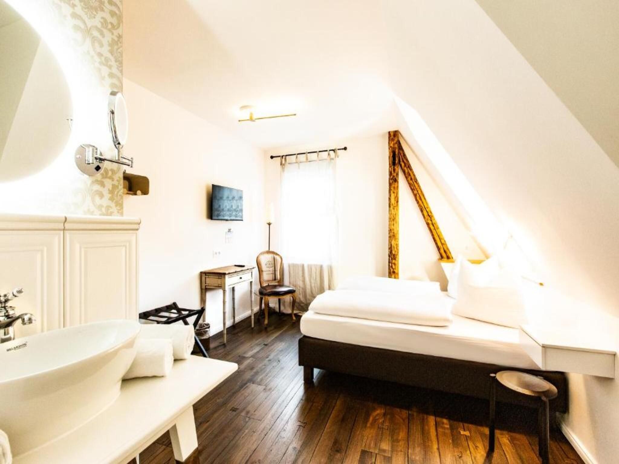 Ein Schlafzimmer mit Bett, Schreibtisch, Stuhl, einem großen Fenster und einem Badezimmer mit Waschbecken und Spiegel.