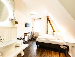 A bedroom with a bed, a desk, a chair, a large window, and a bathroom with a sink and mirror.
