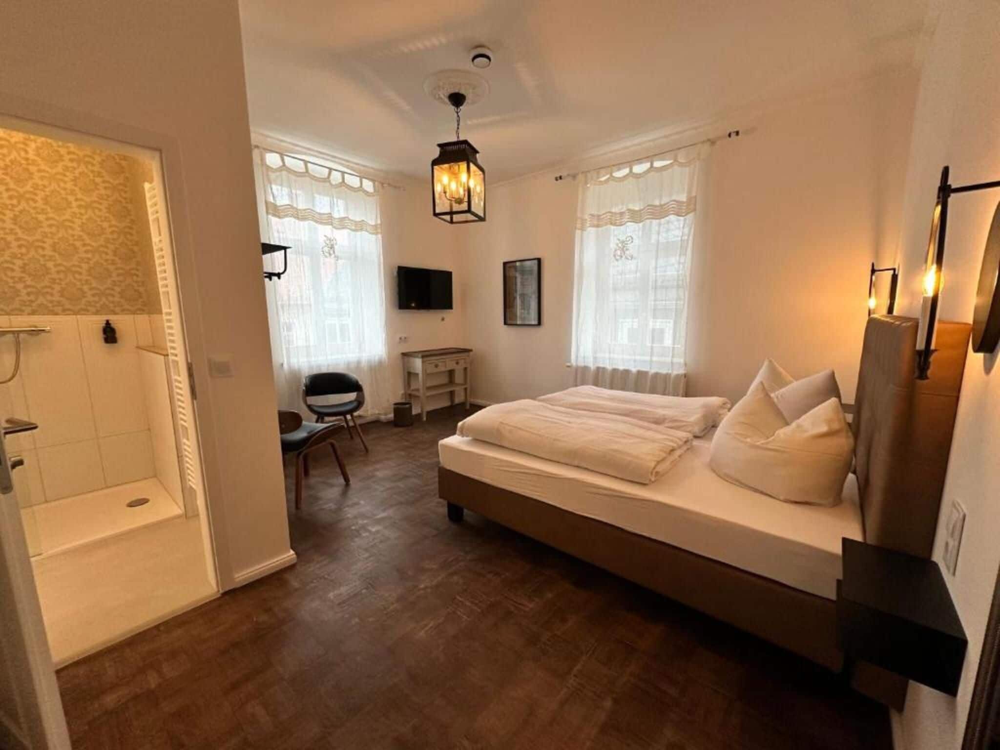 A bedroom with a large bed, a desk, a chair, a television, and a shower area.