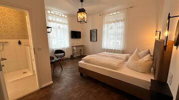 A bedroom with a large bed, a desk, a chair, a television, and a shower area.