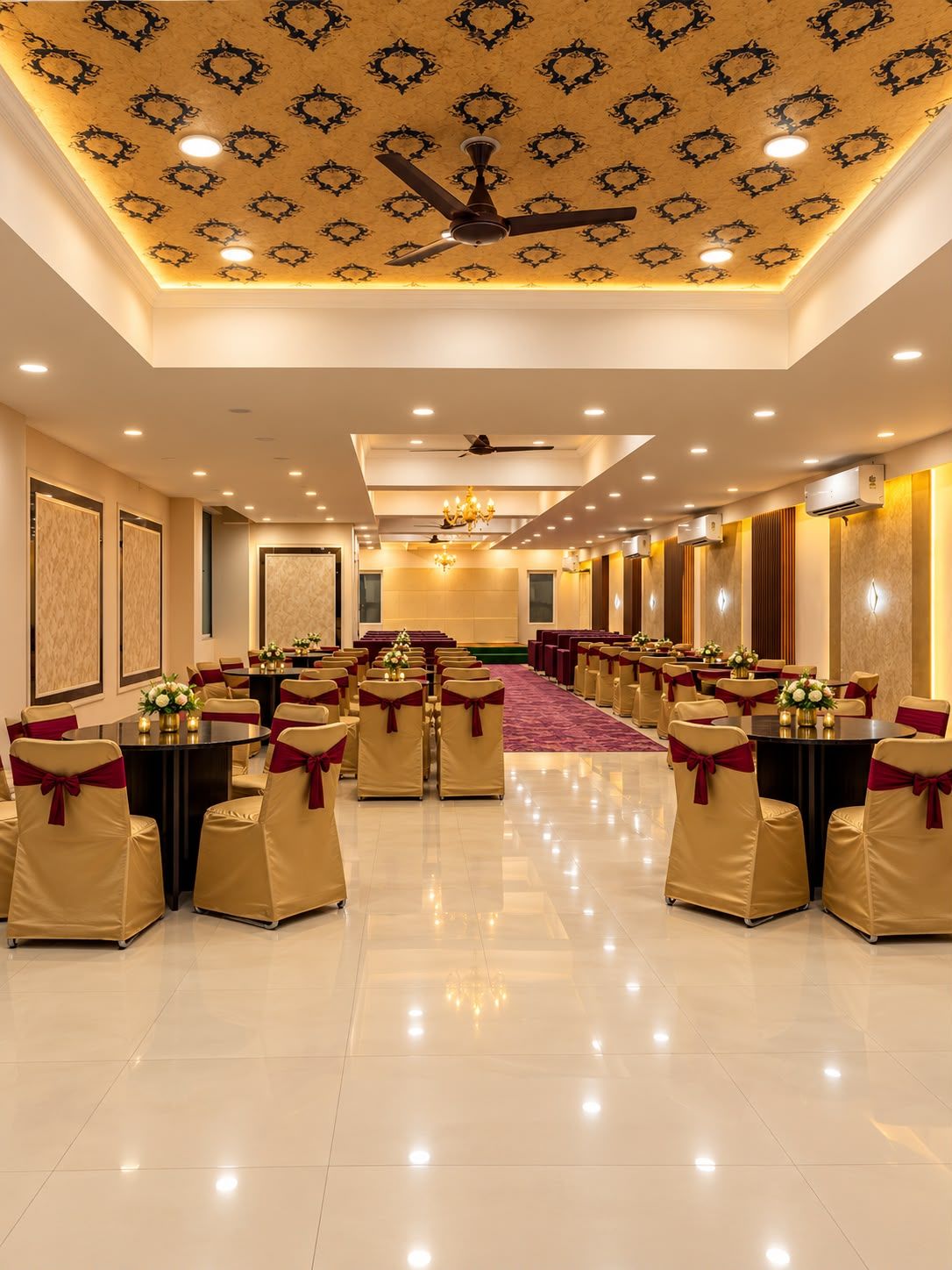 A banquet hall with round tables, chairs with red sashes, a patterned ceiling, and recessed lighting.