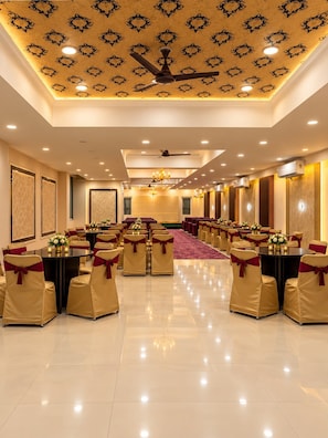 Une salle de banquet avec des tables rondes, des chaises ornées de rubans rouges, un plafond décoré et un éclairage encastré.