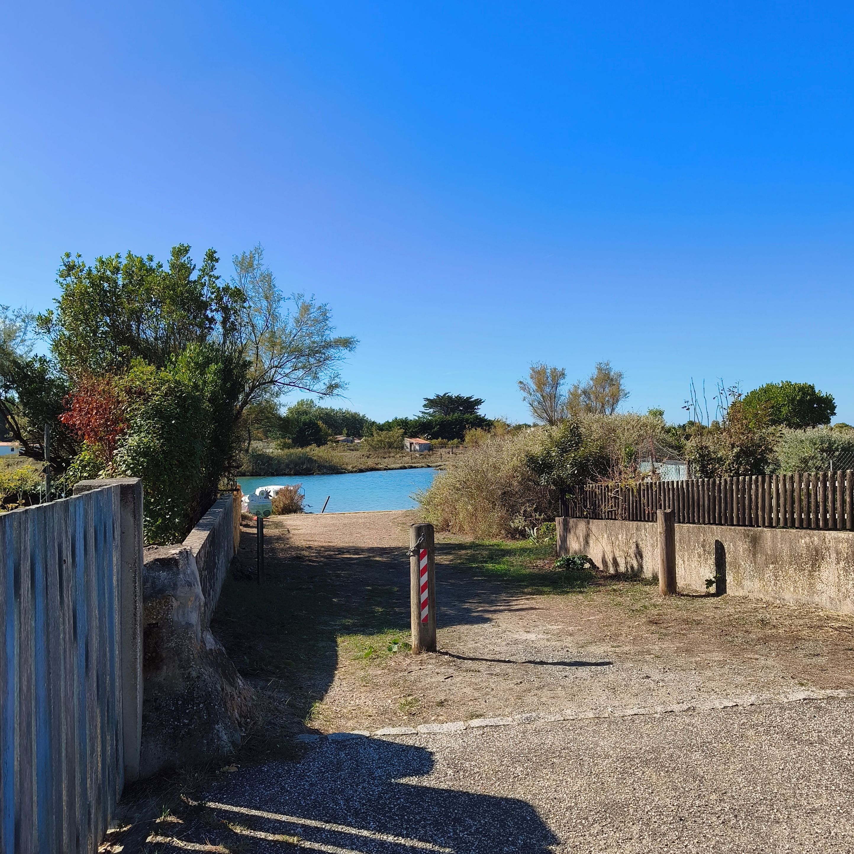 A gravel path leading to a body of water, surrounded by vegetation and a wooden fence.