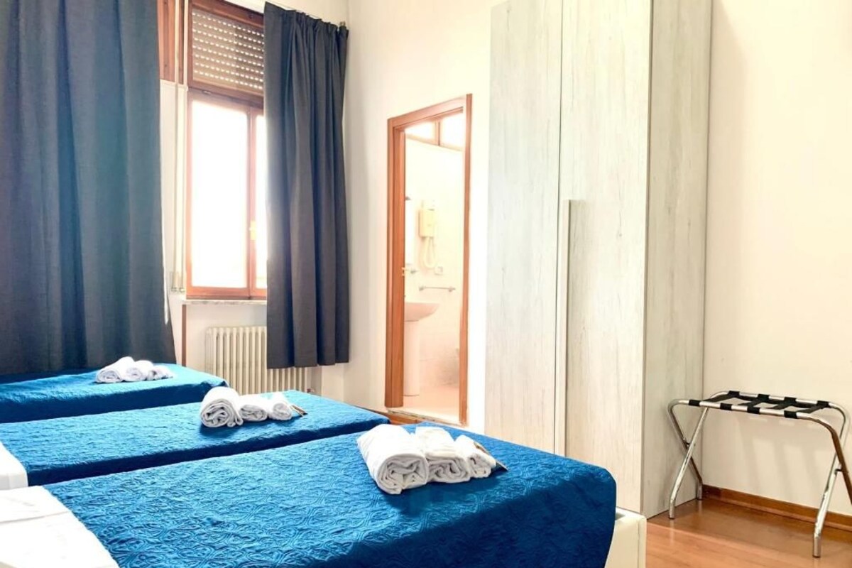 A hotel room with two beds, a radiator, a window with curtains, and a small table.