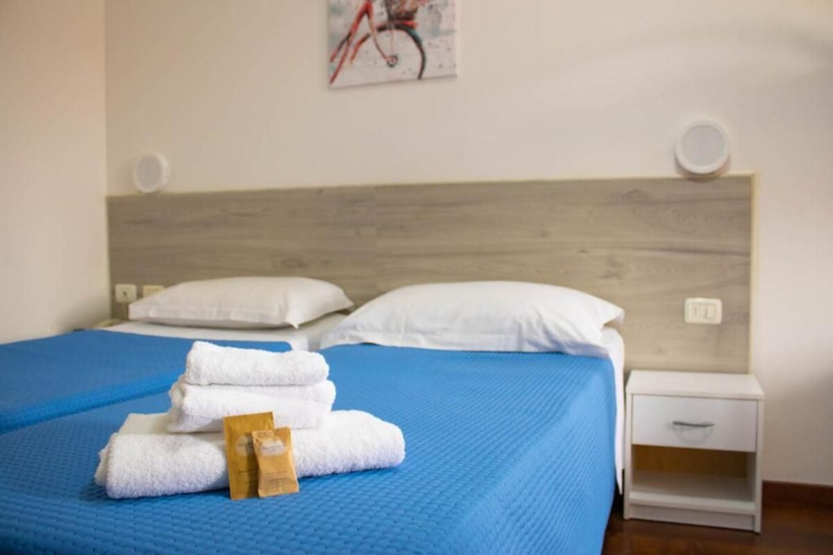 A bed with blue bedding, white pillows, and a stack of white towels. There is a small white bedside table with a drawer.