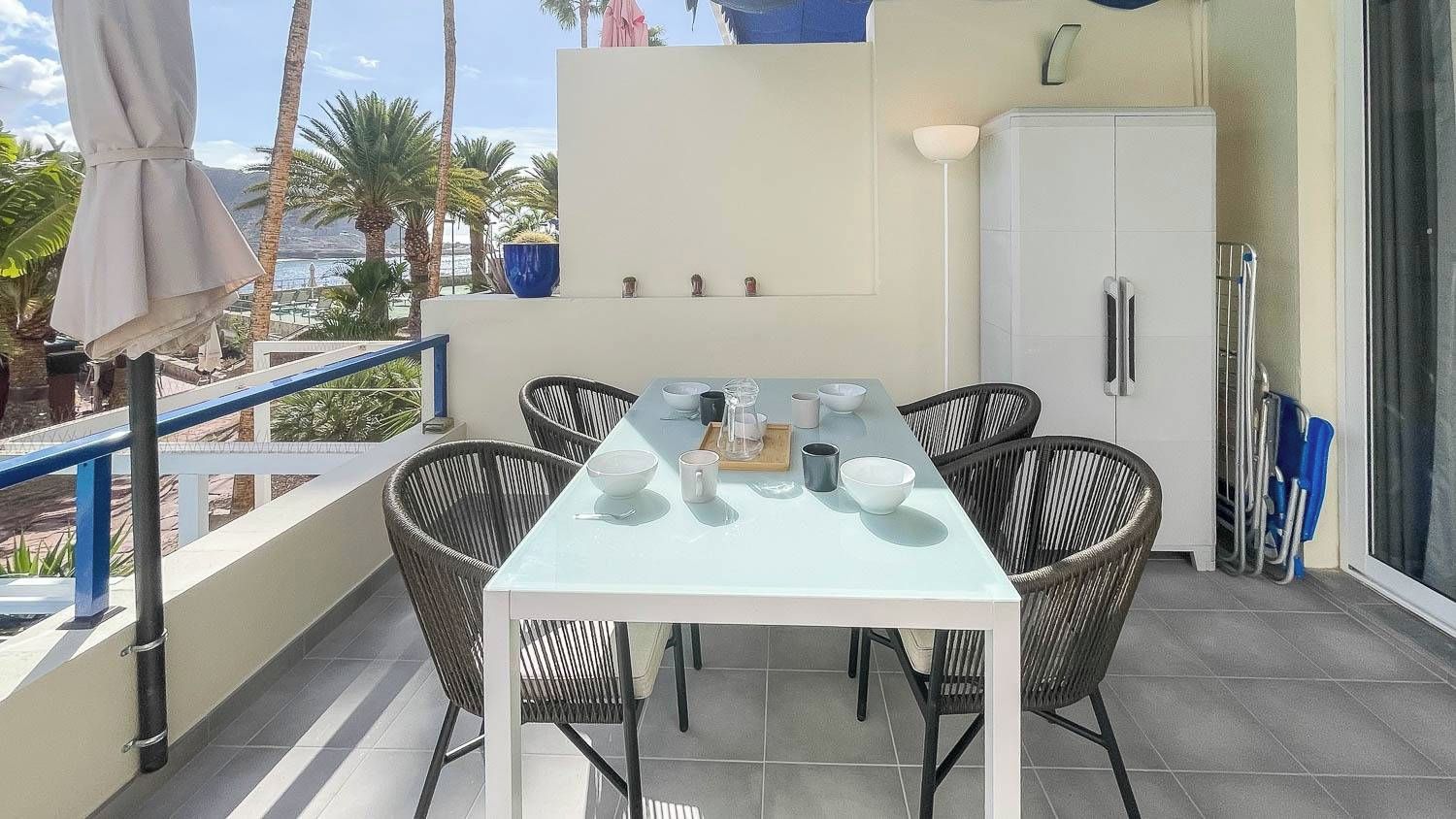 Un balcón con una mesa y sillas, un armario blanco y vistas a palmeras y al mar.