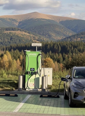 A green electric vehicle charging station with a car connected to it, set against a backdrop of a mountainous landscape.