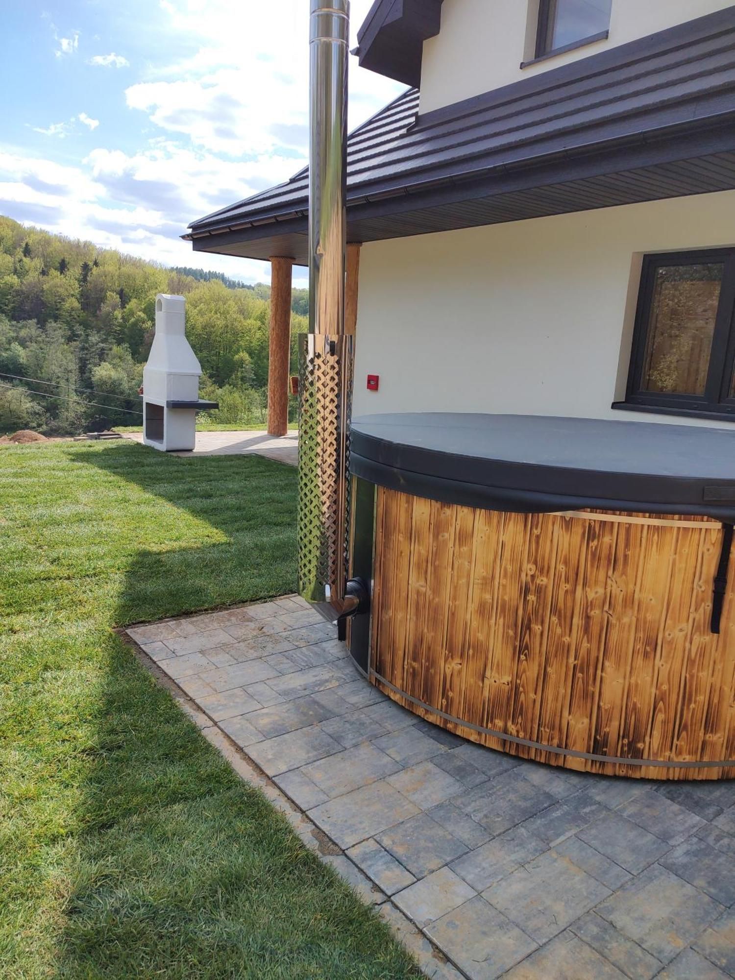 An outdoor bar area with a wooden counter and a grill, set against a house with a sloped roof and a view of a forested area.