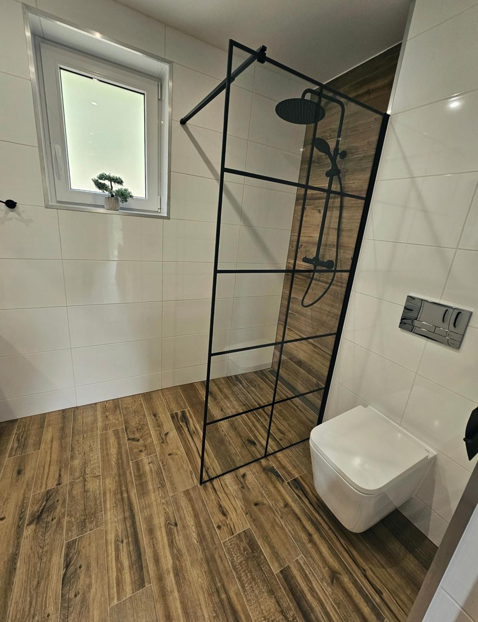 Un baño moderno con suelo de madera, inodoro blanco, cabina de ducha y una ventana con planta en maceta.