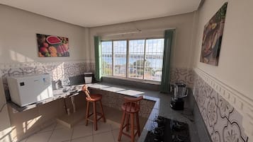 A kitchen with a refrigerator, sink, two stools, a window with a view, and a blender.