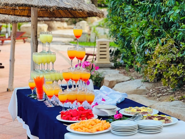 A buffet table with various food items and drinks, set outdoors with a thatched umbrella and greenery in the background.