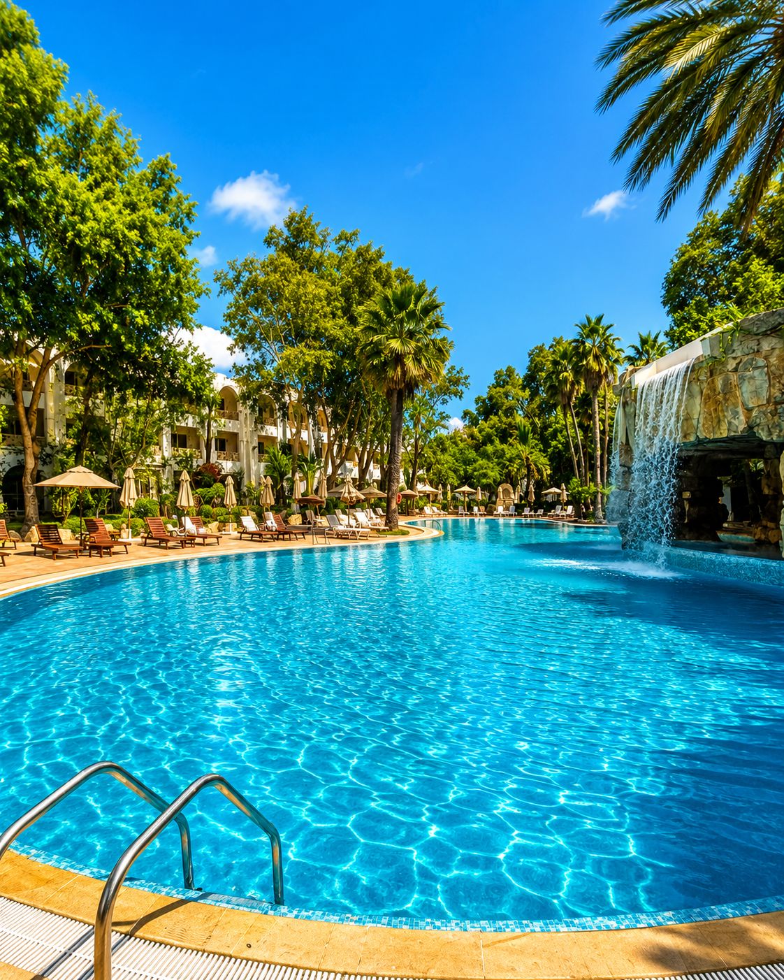 A large outdoor swimming pool with clear blue water, surrounded by palm trees and lounge chairs.
