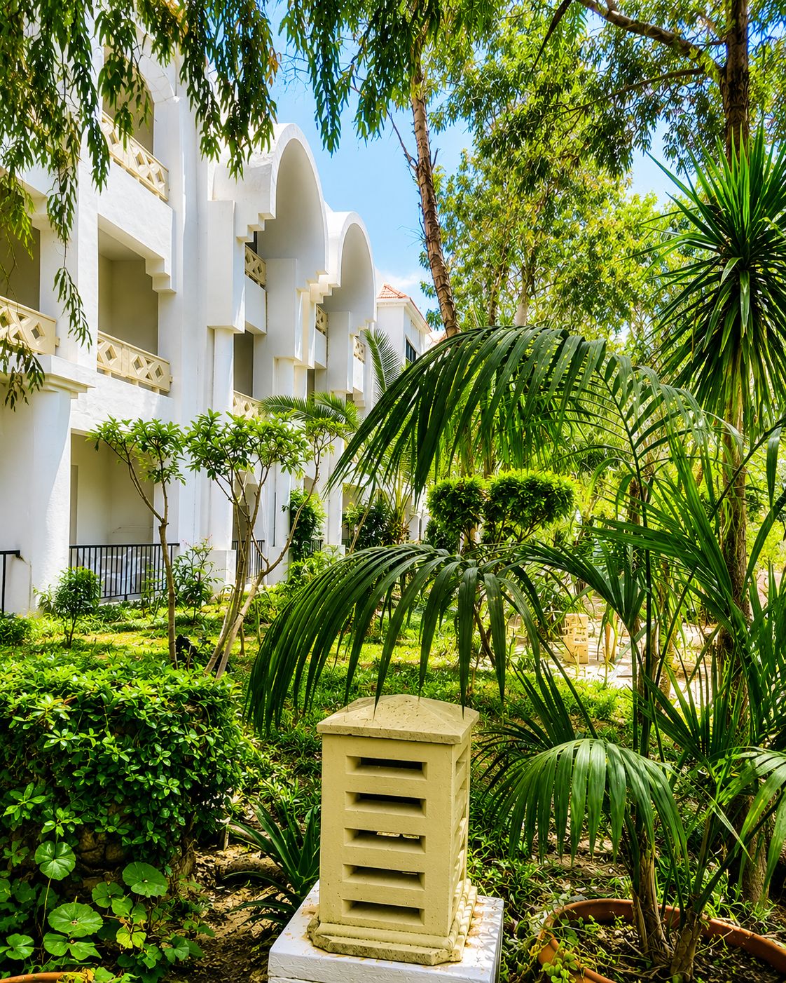 A courtyard with lush greenery, a white building with balconies, and a small beige structure.