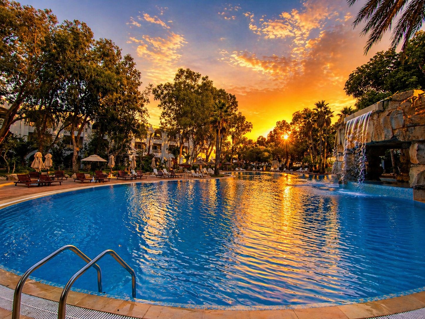 A swimming pool with lounge chairs and umbrellas, surrounded by trees and a sunset.