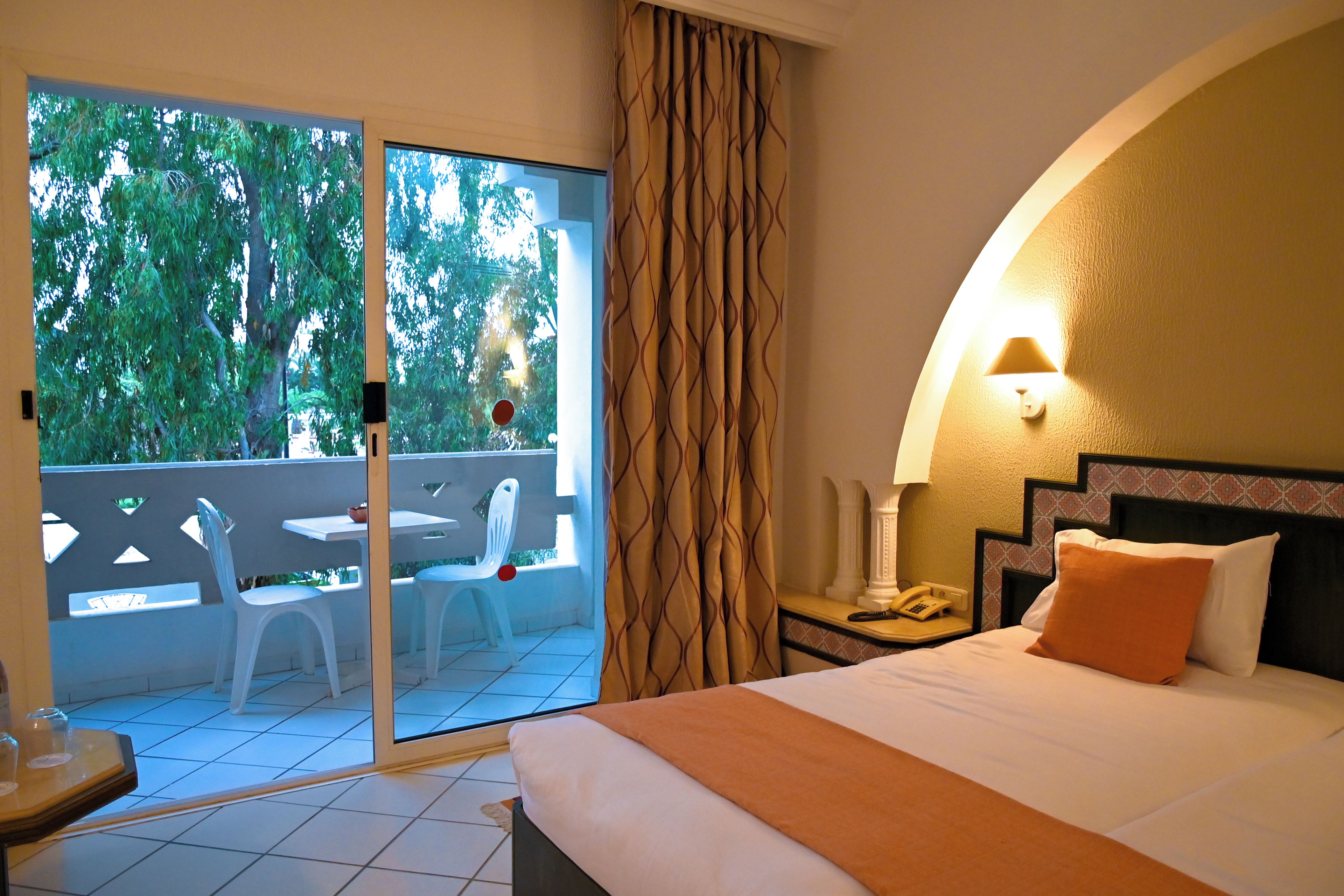 A hotel room with a bed, a bedside table with a lamp, a telephone, and a view of a balcony with chairs and a table.