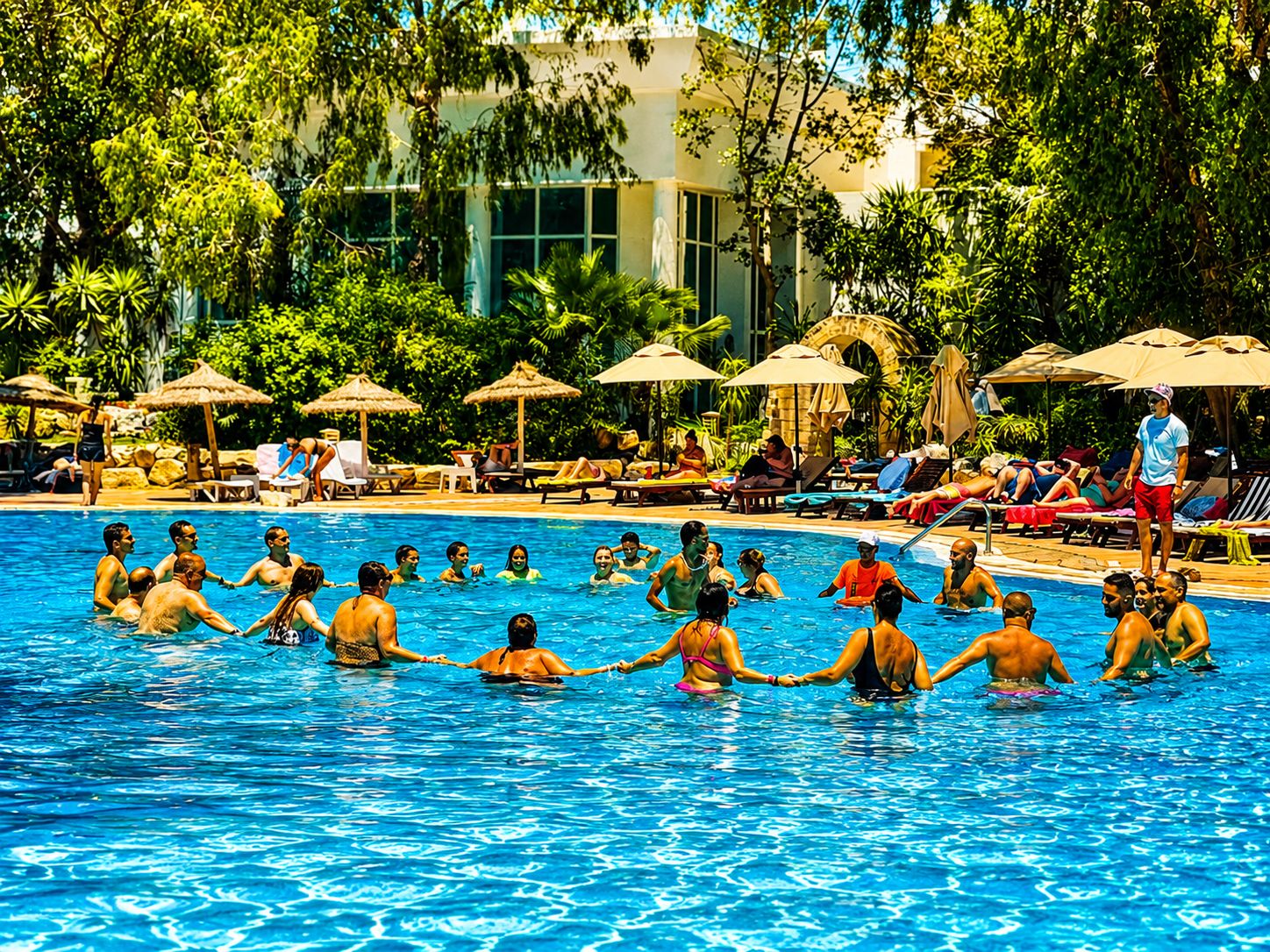 Piscina com pessoas de mãos dadas, rodeada por espreguiçadeiras e guarda-sóis, com um edifício ao fundo.