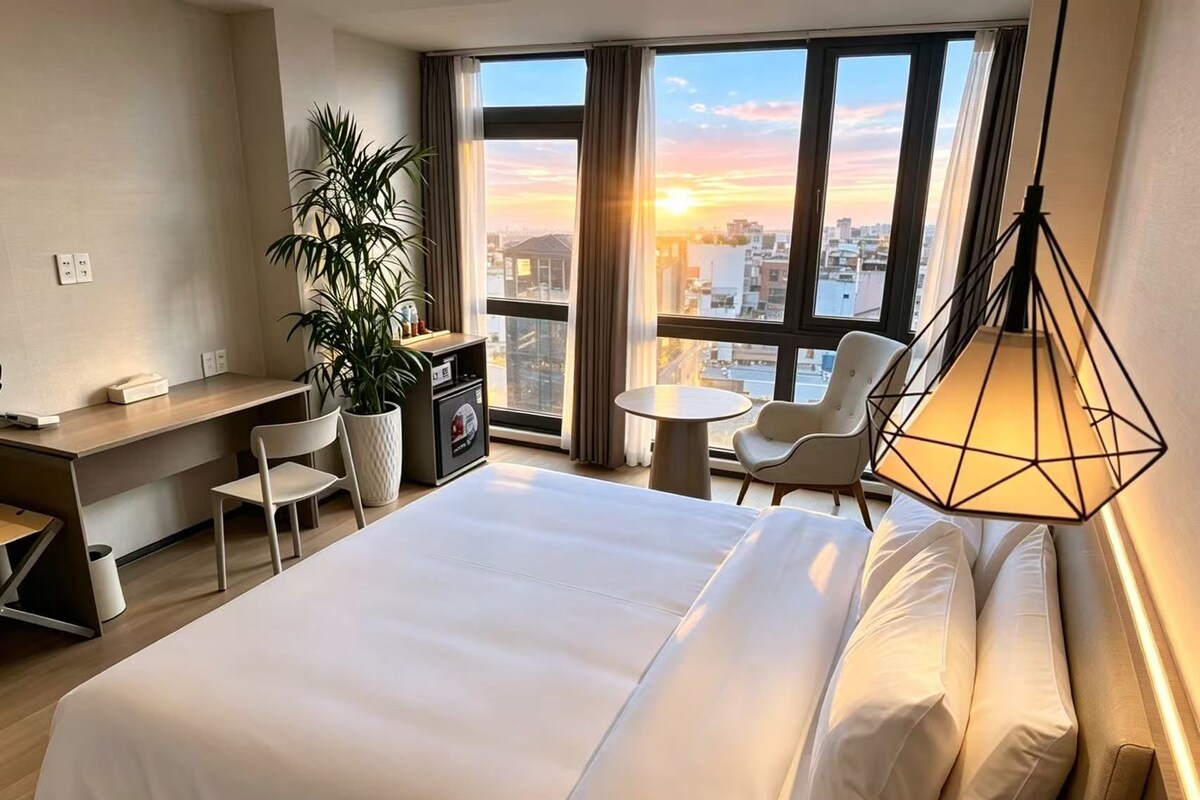 A hotel room with a large bed, a desk, a chair, a small table, and a potted plant.
