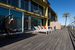 Una terrassa de fusta amb gandules i una cadira penjant, amb vistes a un edifici amb grans finestres i un balcó.